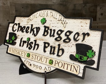 May include: A wooden pub sign with the words "Cheeky Bugger Irish Pub" in black. The sign has a distressed white finish, black border, and shamrock and hat details. It also reads "Strong Ales & Tall Tales" and "Whiskey Stout Poitin".