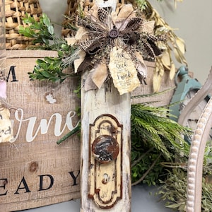 May include: A rustic, decorative wooden door knob plaque with a distressed, off-white finish. It features a metal knob and a decorative floral arrangement with a button. The plaque has a jute rope for hanging and a tag with the text "Ask and it will be given to you; seek and you will find; knock and the door will be opened to you."