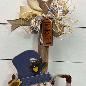 May include: Wooden fall-themed garden tool decoration. Features a snowman face, a blue hat with a black bird, and a tag that says "Hello Fall". The handle is wrapped with twine and adorned with burlap and plaid ribbons.
