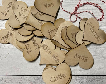 May include: A collection of wooden heart-shaped cutouts with engraved text. The hearts are light brown and feature phrases like "Love," "Kiss Me," and "Yes." A red and white twine is also present, suggesting a craft or decorative item.