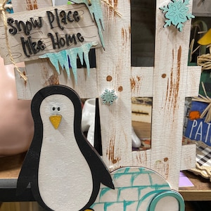 May include: Wooden winter-themed decoration featuring a white picket fence, a penguin, and an igloo. The sign reads "Snow Place Like Home." The penguin is black and white with a yellow beak and feet. The fence has a distressed look with blue icicles and snowflakes.