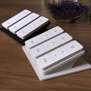 Puede incluir: Mandos a distancia de luz inteligente rectangulares blancos con la palabra "hue" y símbolos de control. Los mandos están sobre soportes blancos y negros, sobre una superficie de madera. En el fondo hay cuencos de cristal con esferas decorativas.