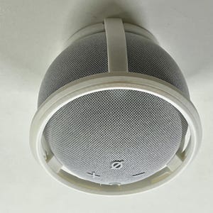 Pode incluir: Suporte de teto branco para um alto-falante inteligente redondo com uma grade de tecido cinza. O suporte tem uma moldura de plástico branco com um design circular.