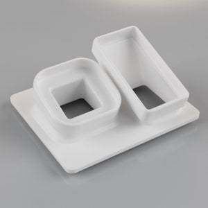 May include: A white ceramic serving dish with a rectangular base. It has two raised compartments: a square with a central opening and a rectangular one. The design is minimalist and modern, suitable for serving appetizers or small portions.