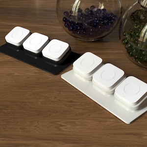 Puede incluir: Dos bases rectangulares, una negra y otra blanca, cada una con tres botones cuadrados blancos con centros circulares. Los botones están dispuestos en fila sobre cada base, presentando un diseño geométrico limpio con un acabado liso y una pequeña luz indicadora.