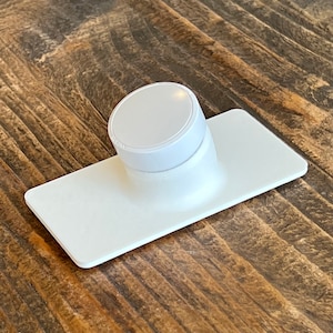 May include: A white, round, magnetic phone holder with a flat base. The holder is designed to attach to a metal surface.