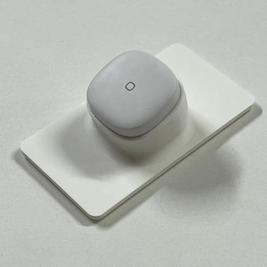 May include: A white, round, wireless charging pad with a small, circular indentation on the top. The charging pad is sitting on a white, rectangular base.