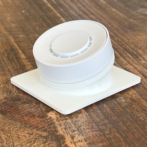 May include: A white round smart home device with a white square base. The device has a circular design with a raised centre and a series of small holes around the edge.