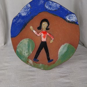 May include: A hand-painted ceramic plate with a whimsical design. The plate features a figure in a red shirt and black pants walking across a brown landscape, with a blue sky and white clouds above. Green accents frame the scene.