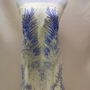 Puede incluir: Tela de tul marfil con bordado de cuentas azules y plateadas. El bordado presenta un diseño floral con hojas delicadas y detalles de cuentas.