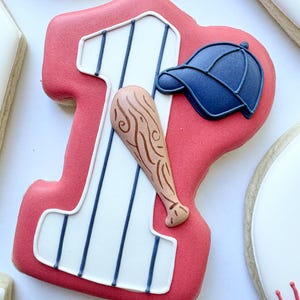 May include: Baseball-themed sugar cookies. A large number one cookie with a baseball bat and cap. Other cookies include a baseball, a baseball glove, and a cookie with the word "YES". The cookies are decorated with red, white, and blue icing.