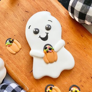 May include: A white ghost cookie with a smiling face and black eyes holds a small orange pumpkin cookie decorated with colorful sprinkles. Three additional pumpkin cookies are on a wooden surface.