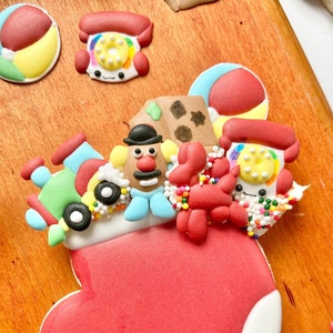 May include: A red and white stocking filled with toy themed cookies. The cookies include a red and yellow ball, a red phone, a brown box, a Mr. Potato Head, a red crab, and a blue train. The cookies are decorated with colourful sprinkles.