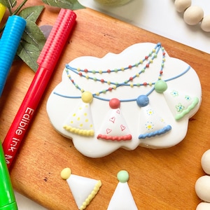 Op de afbeelding: Een witte suikerkoek versierd met een blauwe, groene en gele guirlande van feestmutsen. De koek is gevormd als een wolk en heeft een gegolfde rand. Er liggen ook drie kleinere feestmutskoekjes op een houten oppervlak.