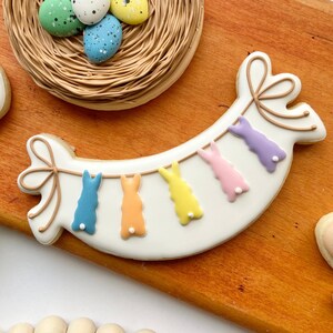 May include: A white cookie decorated with a banner of five pastel coloured bunny rabbits hanging from a string. The banner is on a wooden surface.