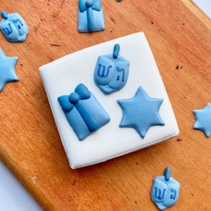 May include: A white square cookie decorated with blue fondant shapes, including stars, dreidels, and gift boxes. The cookie is on a wooden surface with other cookies and decorations.