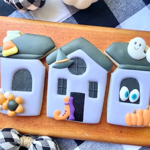 Puede incluir: Tres galletas de azúcar decoradas con forma de casas. La primera casa tiene un techo verde con un maíz dulce en la parte superior, una flor morada y una ventana negra. La segunda casa tiene un techo verde con un sombrero de bruja, una puerta negra y una ventana negra. La tercera casa tiene un techo verde con dos fantasmas blancos, una ventana negra con dos ojos azules y tres calabazas naranjas.