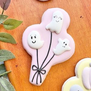Pode incluir: Um biscoito rosa com cobertura de glacê em forma de cacho de balões, decorado com três doces brancos em forma de fantasmas. Os fantasmas têm expressões faciais diferentes. A cobertura preta forma os fios dos balões e um laço. Outros biscoitos com tema de Halloween são visíveis.