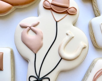 Puede incluir: Una galleta de azúcar decorada con forma de ramo, con un sombrero de vaquero, un pañuelo y una herradura. La galleta está decorada con glaseado marrón claro y negro sobre un fondo blanco. Se ven otras galletas en el fondo.