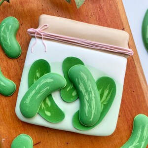 May include: A decorated cookie shaped like a glass jar filled with green pickle slices. The jar has a brown lid with a pink ribbon tied around it. The cookie is on a wooden surface.