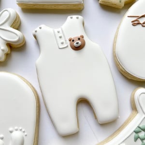 May include: White sugar cookies decorated with white icing and brown bear details. The cookies are shaped like a onesie, a foot print, a rocking horse, and a flower.