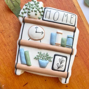 Könnte beinhalten: Ein weißes Keramikregal mit braunem Holzrahmen, verziert mit einer Uhr, Büchern, Pflanzen und einem Schild mit der Aufschrift "HOME".