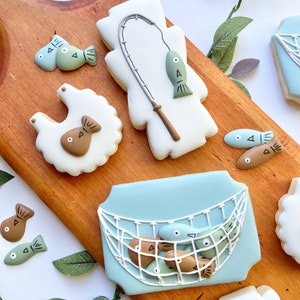 Puede incluir: Un conjunto de seis galletas de azúcar decoradas con temática de pesca. Las galletas están decoradas con glaseado azul, marrón y verde y presentan imágenes de peces, una red de pesca, una caña de pescar y un pelele para bebé.
