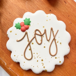 May include: White gingerbread cookie decorated with brown icing in the shape of the word "joy" with a green holly leaf and red berries on top. The cookie is covered in gold sprinkles.
