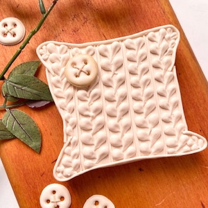 Może przedstawiać: Biała ceramiczna poduszka w kształcie dzianinowego poduszki z guzikiem. Poduszka leży na drewnianej powierzchni z trzema białymi guzikami.