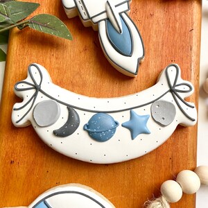 May include: A white sugar cookie decorated with a blue and gray space-themed banner. The banner features a crescent moon, a planet with rings, a star, and two gray moons.