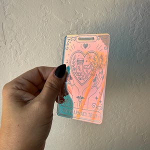 May include: A rectangular iridescent badge holder featuring a tarot-style graphic of two hands holding a heart containing a prescription bottle, mortar and pestle, and the text PHARMACY TECH. The badge has a slot at the top for a lanyard attachment.