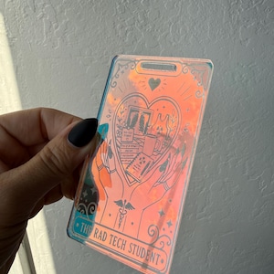 May include: A rectangular iridescent acrylic badge holder featuring a black line art illustration of two hands holding a heart containing a chest X-ray, books, and a mug. The text THE RAD TECH STUDENT is printed at the bottom of the badge.