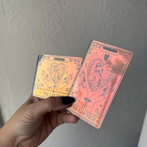 May include: Two rectangular iridescent acrylic badge reels featuring a tarot-inspired design. The graphic depicts hands holding a heart containing a lab technician at a microscope, with the text THE LAB TECH printed at the bottom of each piece.