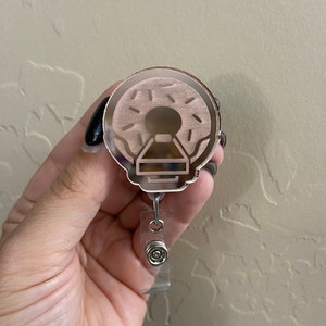 Peut inclure: Un porte-badge en acrylique transparent avec un motif de beigne. Le beigne est brun avec des pépites blanches et un glaçage rose. La bobine est attachée à un cordon rétractable argenté.