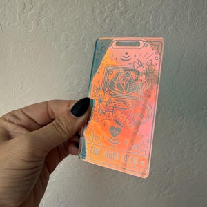 May include: A rectangular iridescent badge holder featuring a holographic finish that shifts between orange and blue tones. The surface is etched with medical illustrations including an ultrasound monitor, a heart, and flowers, with the text THE ECHO TECH.