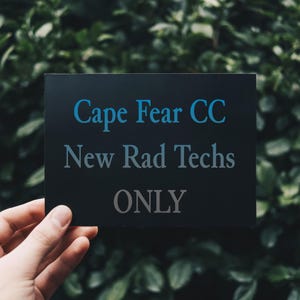 May include: A black rectangular sign with the text "Cape Fear CC" in blue, "New Rad Techs" in gray, and "ONLY" in gray. The sign is held in front of a green leafy background.