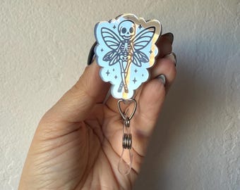May include: A silver badge reel featuring a skeleton fairy design. The fairy has detailed wings and is set against a cloud-shaped background. The reel includes a clear, teardrop-shaped charm and a retractable cord.