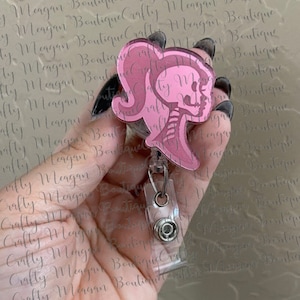 May include: Pink badge reel with a pink silhouette of a woman's profile and a skeleton design. The badge reel has a clear plastic clip and a retractable cord. The text "Meagan Boutique Crafty" is visible in the background.