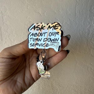 May include: A badge reel with the text "ASK ME ABOUT OUR TURN DOWN SERVICE" in black and gold lettering. The reel has a small charm of a bed with a person in it and a small charm of a cocktail.