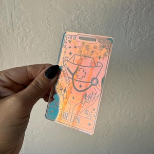 Puede incluir: Un porta credenciales hologr&aacute;fico rectangular con una ilustraci&oacute;n de l&iacute;neas negras de una cofia de enfermer&iacute;a, un estetoscopio, una jeringa y manos. El texto THE LPN est&aacute; impreso en la parte inferior. La superficie refleja tonos naranja, azul y amarillo.