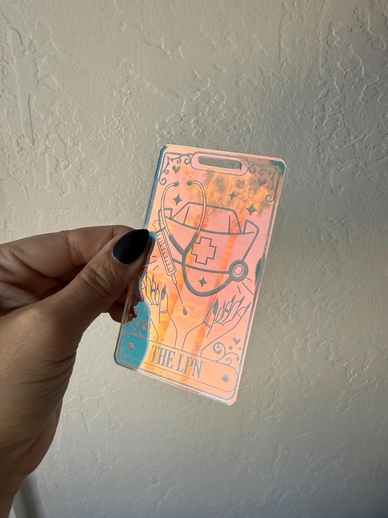 Puede incluir: Un porta credenciales hologr&aacute;fico rectangular con una ilustraci&oacute;n de l&iacute;neas negras de una cofia de enfermer&iacute;a, un estetoscopio, una jeringa y manos. El texto THE LPN est&aacute; impreso en la parte inferior. La superficie refleja tonos naranja, azul y amarillo.