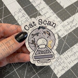 May include: A white sticker with a gray circle and the text "Cat Scan" in black. Inside the circle are images of cats in various poses. Below the circle is a gray rectangle with the text "Tech" in white.