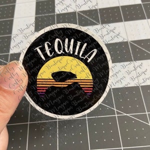 Peut inclure: Un autocollant rond noir et blanc avec le mot "Tequila" en texte blanc. L'autocollant a un graphique de coucher de soleil avec une silhouette d'une personne buvant dans un verre.