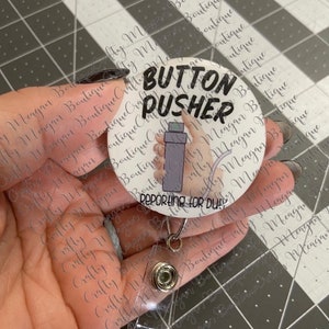 May include: White round button with black text that reads "Button Pusher Reporting for Duty". The button has a cartoon image of a hand holding a bottle of hand sanitizer. The button is attached to a clear plastic retractable badge holder.