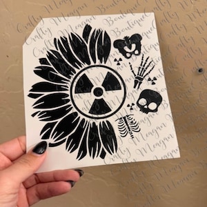 May include: A black and white sticker featuring a sunflower design with a radiation symbol at the center. Additional elements include a skull, skeleton hand, rib cage, and pelvis. The sticker is held by a hand.