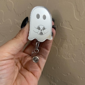 May include: A silver ghost-shaped badge holder with a small, silver radiation symbol inside. The ghost is holding the symbol in its hands. The badge holder is attached to a clear plastic retractable badge reel.