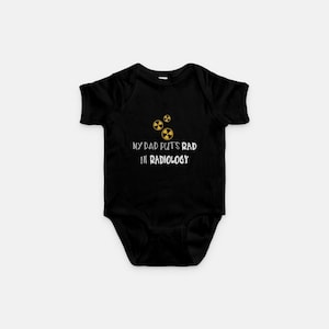 May include: Black baby bodysuit with the text "MY DAD PUTS RAD IN RADIOLOGY" in white. The design includes three yellow radiation symbols. The bodysuit is short-sleeved and has a simple, clean design.
