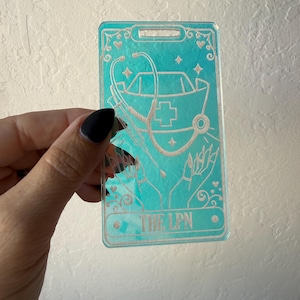 Puede incluir: Un porta credenciales rectangular de acr&iacute;lico transl&uacute;cido color verde azulado con un dise&ntilde;o grabado de una cofia de enfermer&iacute;a, un estetoscopio y una jeringa sostenidos por dos manos. El texto THE LPN est&aacute; grabado abajo con estrellas y corazones.