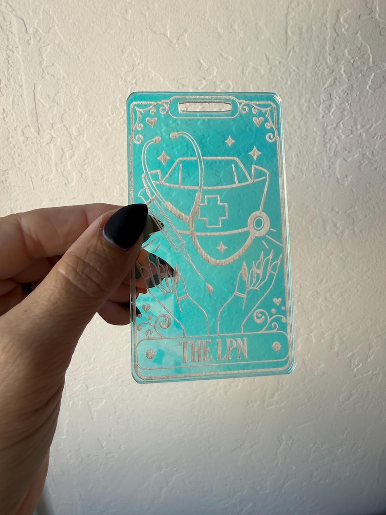 Puede incluir: Un porta credenciales rectangular de acr&iacute;lico transl&uacute;cido color verde azulado con un dise&ntilde;o grabado de una cofia de enfermer&iacute;a, un estetoscopio y una jeringa sostenidos por dos manos. El texto THE LPN est&aacute; grabado abajo con estrellas y corazones.