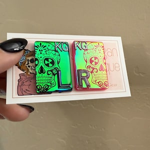 May include: Two enamel pins with a skull design and the letters "KG" and "LR" on a white background. The pins are iridescent and have a green and pink color scheme.